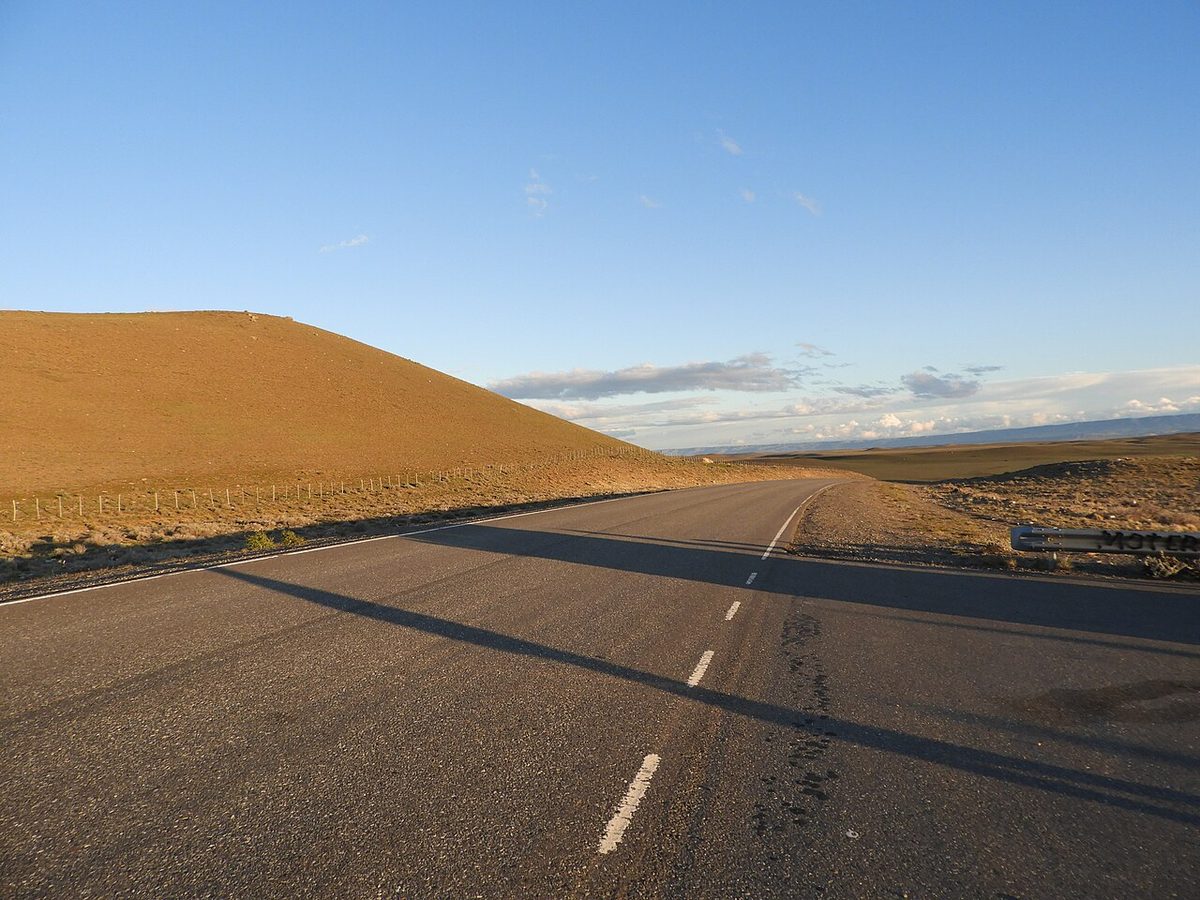 Paved Ruta 40 sweeping past a bare, sunlit hillside on the Santa Cruz steppe
