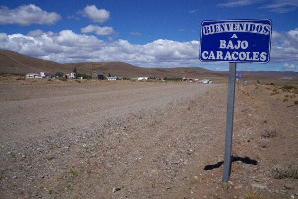 The gravel shoulder of Ruta 40 at the Bajo Caracoles turn-off, with the hamlet's welcome sign and a few buildings on the steppe