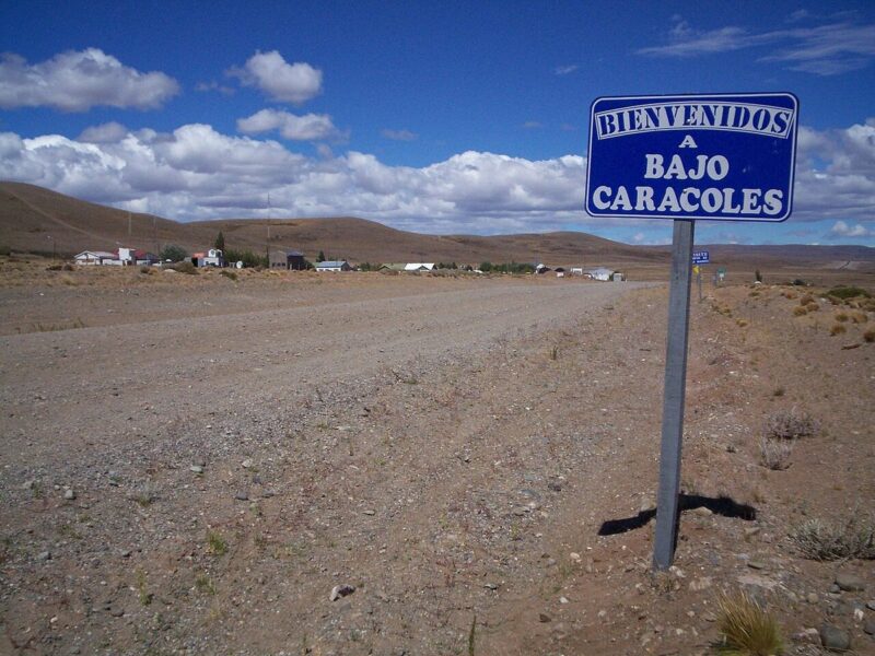 The gravel shoulder of Ruta 40 at the Bajo Caracoles turn-off, with the hamlet's welcome sign and a few buildings on the steppe