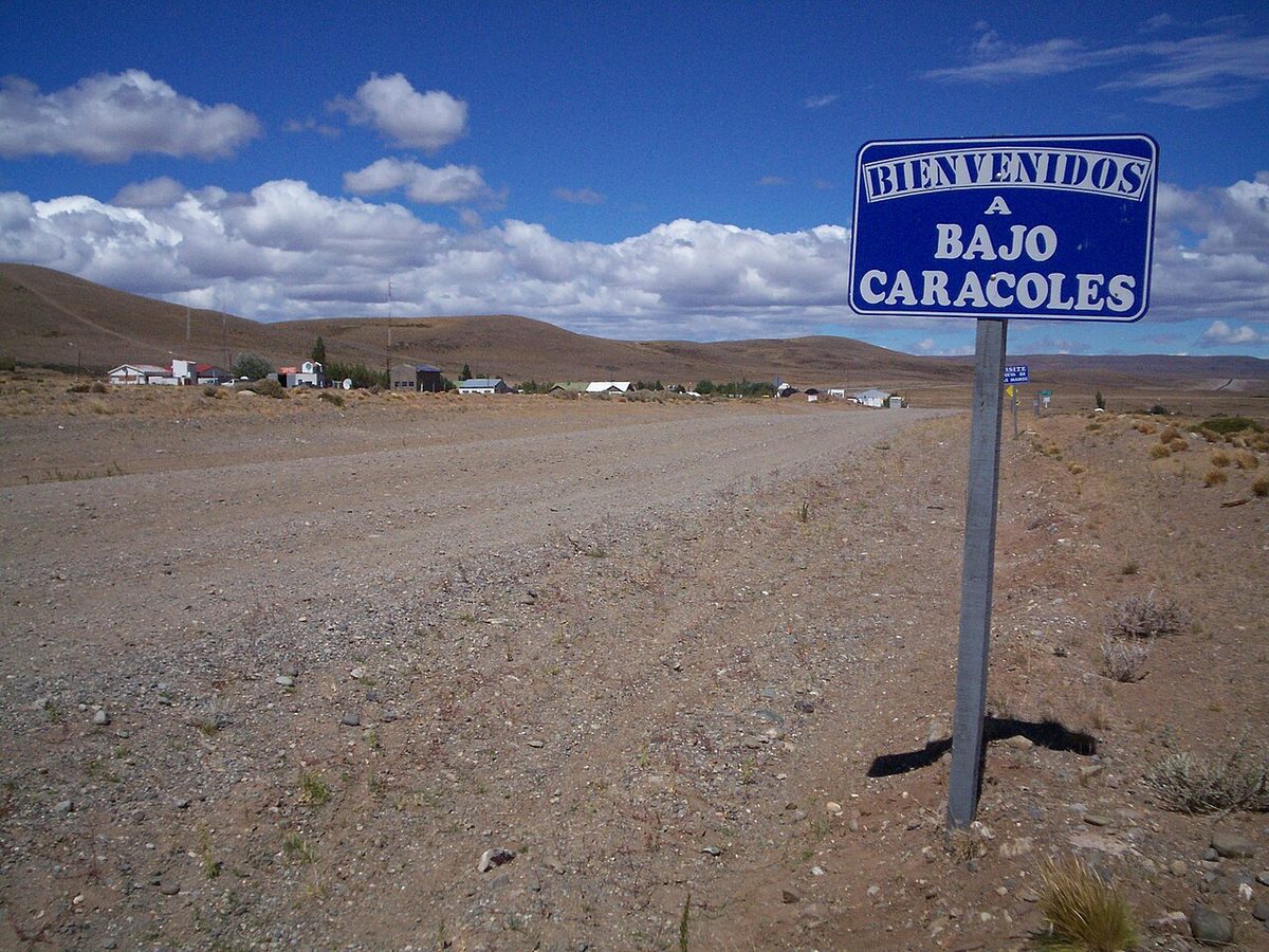 The gravel shoulder of Ruta 40 at the Bajo Caracoles turn-off, with the hamlet's welcome sign and a few buildings on the steppe